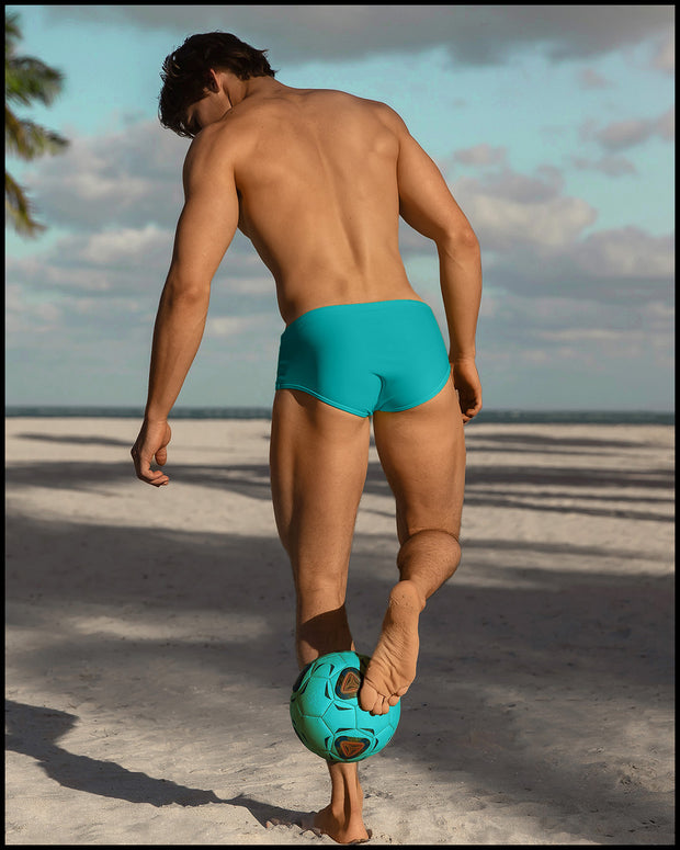 Male model outdoors at the beach playing volleyball wearing the CERULEAN a teal green/blue color Swim Sunga by BANG! Clothes.