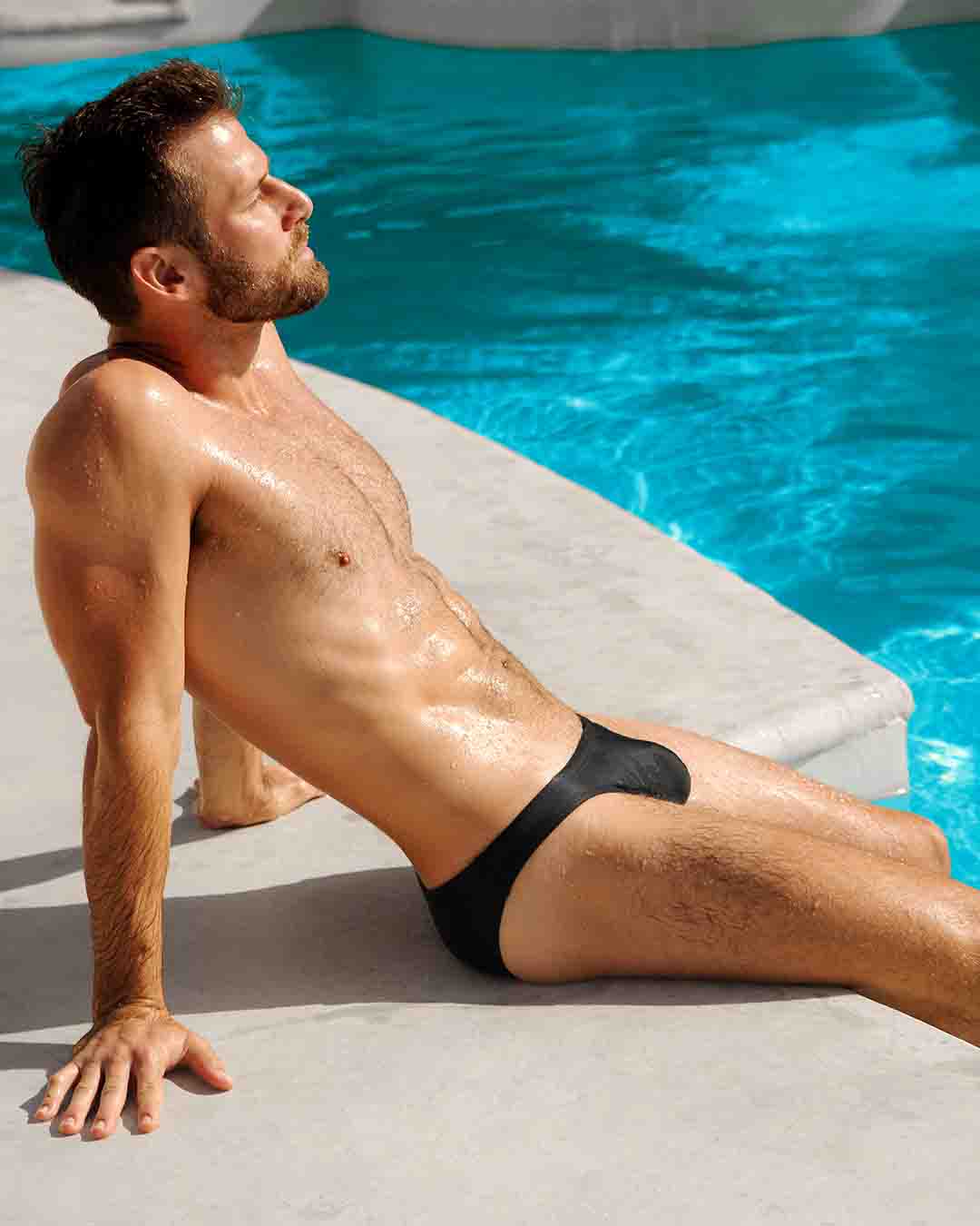 Male model sitting poolside wearing a Swim Sunga of the new JET BLACK dark color men's swimsuit by DC2 Miami for BANG menswear Miami.