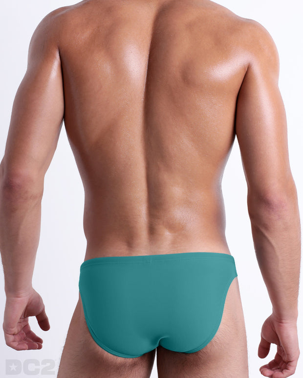 Back view of male model wearing the ATLANTIS TEAL beach mini-briefs for men by BANG! Miami in a solid light teal color.