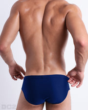 Back view of male model wearing the BLUE BY THE OCEAN beach briefs for men by BANG! Miami in a solid dark blue color.