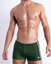 Front view of model wearing the BRAVE GREEN men’s bikini-style Swim Trunks, premium swimwear in a solid dark green color with white and orange stripes on the sides for men. These high-quality swimwear are by DC2, a men’s beachwear brand from Miami.