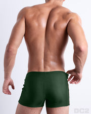 Back view of male model wearing the BRAVE GREEN beach swimming bottoms for men in a solid pine green color with side orange and white stripes, designed by DC2.