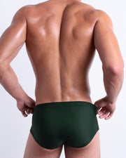 Back view of a model wearing CASINO ROYALE (GREEN) men’s beach sexy swim sunga in dark green color. Inspired by actor Daniel Craig's iconic blue swim trunks worn in the 2006 film Casino Royale, is designed by BANG! Clothes in Miami.