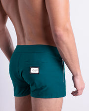 Male model wearing men’s CHIC TEAL Beach Shorts swimsuit in a dark aqua green blue color, complete the back zippered pocket, made by DC2 a capsule brand by BANG! Clothes in Miami.