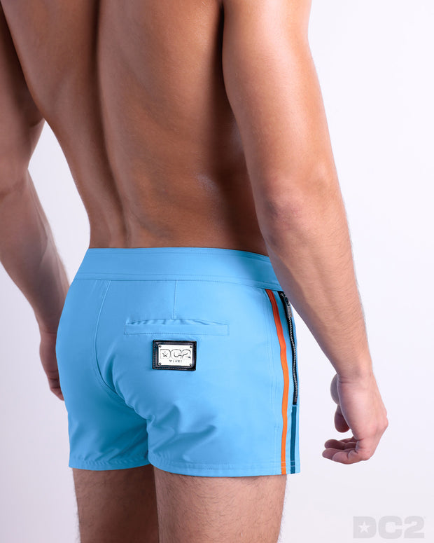 Back view of a male model wearing men’s Summer COASTAL BLUE Beach Shorts in a solid light blue color with orange and dark blue side stripes, complete the back pockets, made by DC2 a capsule brand by BANG! Clothes in Miami.
