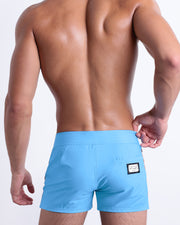 Back view of a male model wearing men’s Summer COASTAL BLUE Beach Shorts in a solid light blue color with orange and dark blue side stripes, complete the back pockets, made by DC2 a capsule brand by BANG! Clothes in Miami.