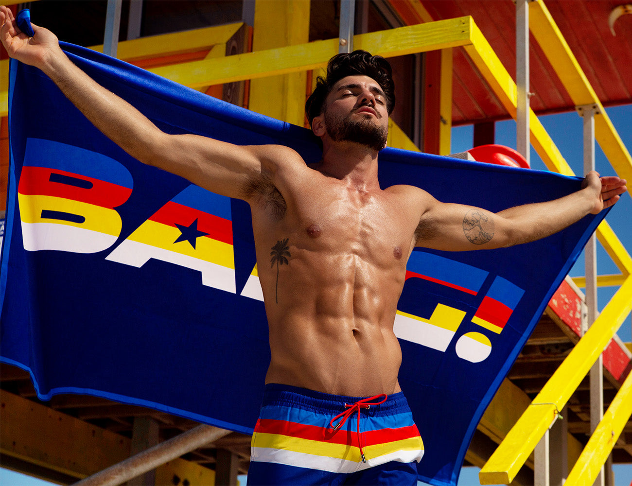 Male model at the beach wearing the STRIPE’A’POSE Show Shorts and holding the matching light-weight & portable beach towel.