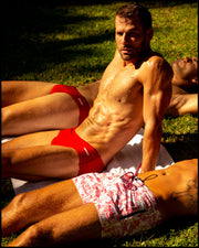Male models laying down outdoors in their BANG! Clothes men’s swimsuit perfect for beach sun tanning activity. The model on the far left is wearing the MAJESTIC RED Swim Brief a bright solid red color. The model in the middle is wearing the MAJESTIC RED Swim Sunga in a true Baywatch red color. Model on the bottom is wearing the TOILE DE MIAMI (RED) Flex Shorts.