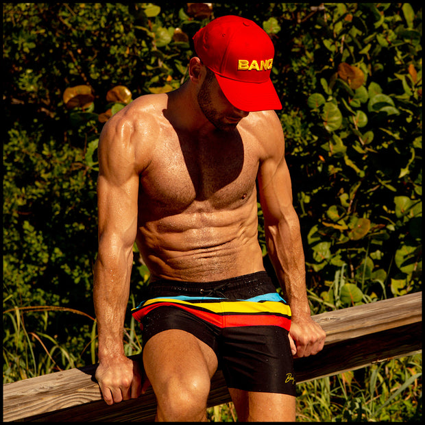 Male model sitting down wearing the BIONIC STRIPES Resort Shorts and the HIGH RED baseball cap by BANG! Clothes at the beach. 