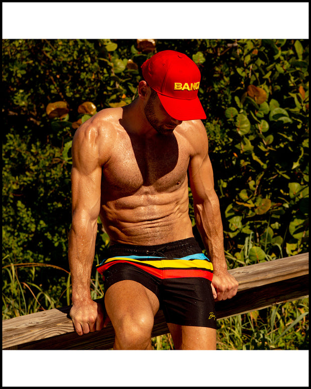 Male model sitting down wearing the BIONIC STRIPES Resort Shorts and the HIGH RED baseball cap by BANG! Clothes at the beach. 
