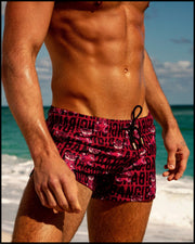 A male model is at the beach, showcasing SHOW SHORTS swimwear for men in STARSTRUCK. These shorts feature a red berry color with black BANG! Typography print and tiger pop art.