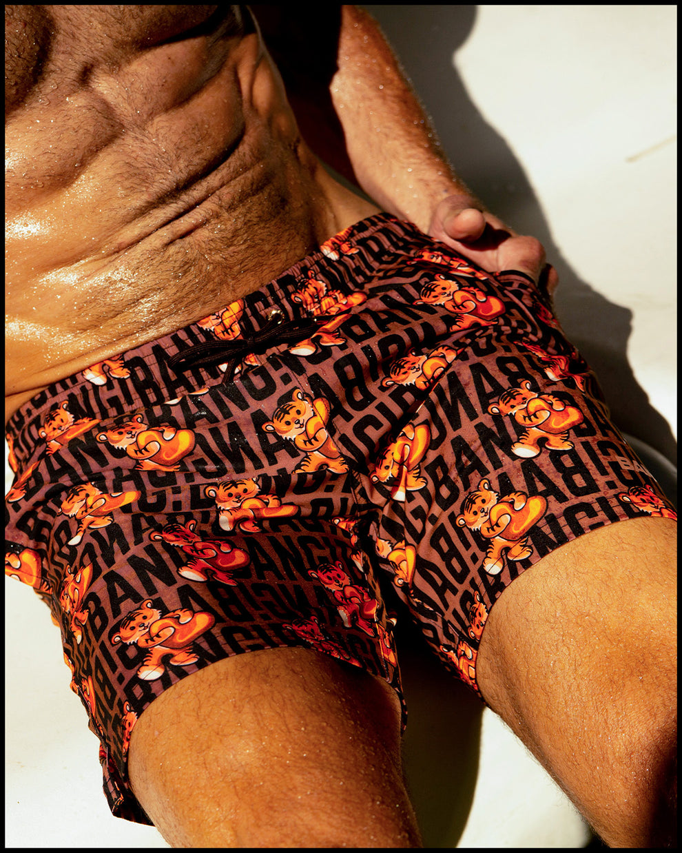 A male model is sitting down, showcasing the back of the "SHOW SHORTS" swimwear for men from TIGER HEARTS. These shorts feature a pop art design of brown and orange tigers by BANG! Clothes.