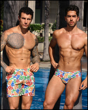 Male models by the pool wearing the VIA POSTAL Series featuring postage stamps of Miami beach Sun, Fun, Sand, Sea, flamingo, dolphin, jet ski, lifeguard tower graphic by Miami based BANG! Miami brand of men's beachwear. Model on the left is VIA POSTAL Resort Shorts and model on the right is wearing the matching VIA POSTAL Swim Brief.