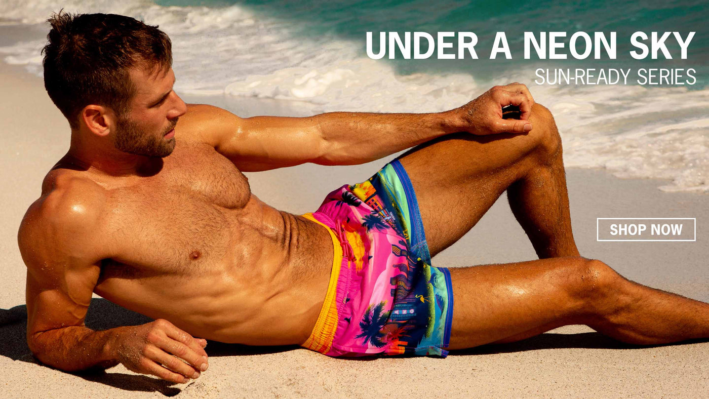 The New UNDER A NEON SKY men's beachwear series by BANG!® Miami. Featuring graphics of miami and south beach in neon pink and orange. The collection includes men's beach shorts and poolside style swimsuits with summer-ready colors to wear under sunny skies.