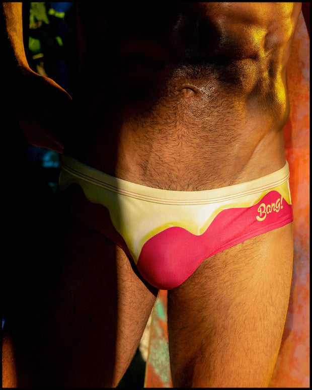 Male model outdoors wearing the MY MILKSHAKE Swim Brief bikini-style bottoms in a bright pink color featuring yellow melting ice cream print by BANG! Clothes the official brand of men’s swimwear based in Miami, FL.