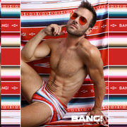 Male model laying down wearing the FAWCETT SARAPE Swim Sunga featuring red, blue, purple, and white colors inspired by the famous 1976 swimsuit poster of Farrah Fawcett. This swimsuit is by BANG! Clothes official brand of men’s high-quality beachwear. 