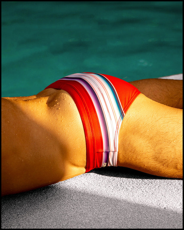 Male model laying down wearing the FAWCETT SARAPE Swim Brief featuring red, blue, purple, and white colors inspired by the famous 1976 swimsuit poster of Farrah Fawcett. This swimsuit is by BANG! Clothes official brand of men’s high-quality beachwear. 