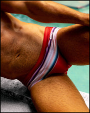 Male model wearing the FAWCETT SARAPE Swim Brief featuring red, blue, purple, and white colors inspired by the famous 1976 swimsuit poster of Farrah Fawcett. This swimsuit is by BANG! Clothes official brand of men’s high-quality beachwear. 