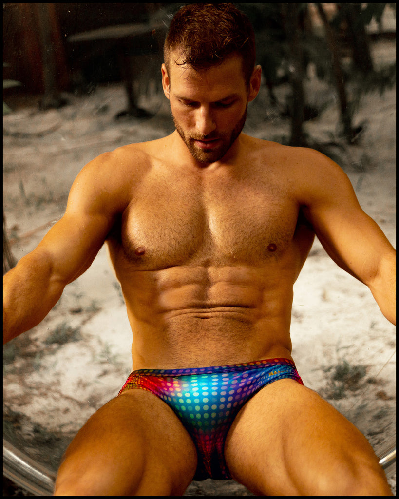 Male model sitting down wearing the CONFESSIONA ON A SAND FLOOR Swim Brief in multiple colors Disco Ball design by BANG! Clothes, the official brand of men’s swimwear.