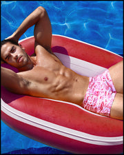 Male model in the pool wearing the TOILE DE MIAMI (RED) Swim Trunks in white with bright red toile jouy art by BANG! Clothes, the official brand of men’s swimwear.
