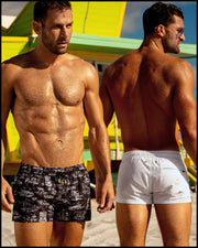 Two male models are pictured at the beach, sporting stylish men’s beach shorts from BANG! Clothes. The brand is Miami-based and specializes in men’s swimwear. On the left, the model is wearing the TOILE DE MIAMI (BLACK) Show Shorts, while the model on the right is wearing the WHITE PARTY Mini Shorts.