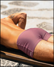Sexy male model at the beach in Brazil wearing the BUST A MAUVE Swim Trunks men’s swimwear in a light mauve color with the official logo of BANG! Brand in white.