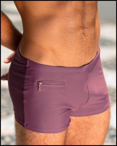 Sexy male model at the beach in Brazil wearing the BUST A MAUVE Swim Trunks men’s swimwear in a light mauve color with the official logo of BANG! Brand in white.