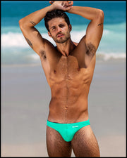 Sexy male model wearing the AQUA GLOW Swim Mini Brief men’s swim European bikini in an aquamarine color made with Italian-made Vita By Carvico Econyl Nylon with the official logo of BANG! Brand in white.