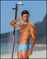 Sexy male model at the beach in Brazil wearing the MAGNET BLUE Swim Sunga men’s swim European bikini in a light blue color made with Italian-made Vita By Carvico Econyl Nylon with the official logo of BANG! Brand in white.