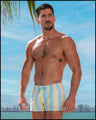 A sexy male model at the beach wearing THE KEN (MIAMI EDITION) Show Shorts. These swimming trunks are inspired by the Barbie movie and the SoBe/Art-Deco style, featuring pastel colors with yellow and blue stripes, reminiscent of the styles worn by Ryan Gosling as Ken in the Barbie movie. The beach shorts are designed by BANG! Clothes, a men’s beachwear brand based in Miami.