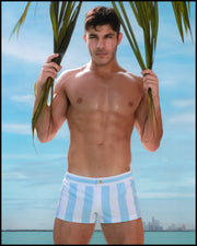 At the beach, a male model is seen wearing THE KEN (MYKONOS EDITION) Mini Shorts. This swimsuit is inspired by Santorini, Greece and features pastel colors with white and blue stripes, similar to the styles worn by Ryan Gosling as Ken in the Barbie movie. The beach shorts are designed by BANG! Clothes, a men’s beachwear brand based in Miami.