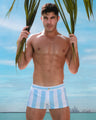 At the beach, a male model is seen wearing THE KEN (MYKONOS EDITION) Mini Shorts. This swimsuit is inspired by Santorini, Greece and features pastel colors with white and blue stripes, similar to the styles worn by Ryan Gosling as Ken in the Barbie movie. The beach shorts are designed by BANG! Clothes, a men’s beachwear brand based in Miami.