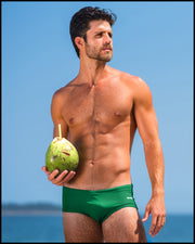 Sexy male model at the beach holding a coconut wearing a men’s Brazilian style swimsuit in GREEN RUSH an emerald green color made with Italian-made Vita By Carvico Econyl Nylon by the Bang! Menswear brand from Miami.