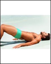 At the beach, a male model is seen wearing BANG! AQUA GLOW Brazilian Swim Sunga in a bold aqua color.