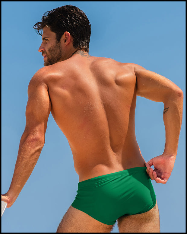 Sexy male model at the beach wearing a men’s Brazilian style swimsuit in GREEN RUSH an emerald green color made with Italian-made Vita By Carvico Econyl Nylon by the Bang! Menswear brand from Miami.