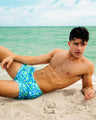 At the beach, a male model is wearing the CAMO POP (BLUE/GREEN) Show Shorts. The swimsuit features a bright sky blue and green neon camo print designed by BANG! Clothes in Miami.