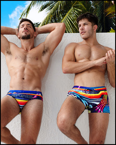 Two male models are seen outdoors leaning against a wall. The model on the left is wearing the POOL POSITION Swim Brief, while the one on the right is wearing the POOL POSITION Show Shorts. This Summer swimsuit for the beach boasts a combination of blue, red, yellow, and orange shades, inspired by the iconography of racing car graphics. The high-quality design of this swimwear pays homage to motorsports and Grand Prix racing, and it is designed by BANG! Clothes in Miami.
