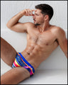 Male model sitting down wearing the POOL POSITION Swim Brief. This Summer swim brief swimsuit for the beach includes shades of blue, red, yellow, and orange - inspired by the iconography of racing car graphics. This high-quality swimwear design pays homage to motorsports and Grand Prix racing, designed by BANG! Clothes in Miami.