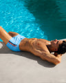 A male model is seen by the pool wearing the Coastal Blue Mini Shorts by DC2, a Miami-based capsule brand from BANG! These beach shorts are light blue with two stripes on the side. One stripe is bright orange, while the other is navy.