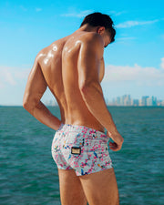 This image features a model wearing CLOSE TO YOU Mini Shorts, with the Miami skyline in the backdrop. The beach shorts have an exotic bird design in light blue color with pink flowers, designed for men. These premium quality swimwear bottoms are by DC2, a popular men’s beachwear brand based in Miami.