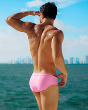 Male model posing outdoors with the Miami skyline in the background in his vibrant PINKTYQUE Swim Brief, a bright pink design by DC2, a men’s beachwear brand from Miami.