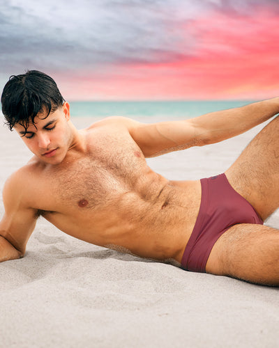 A male model is lying on the beach wearing the SUN-KISSED RED Swim Brief by DC2. This swimsuit comes in a light berry color inspired by Miami’s beautiful sunsets, making it perfect for any summer vacation.