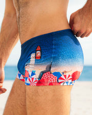 This is a side view of a male model wearing the UNDER MY UMBRELLA Swim Trunks for men at the beach. These swim trunks are premium quality and feature an ombre print of a Miami beach scene. They are designed by DC2, a men's beachwear brand based in Miami.