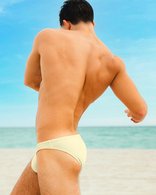 A muscular male model in a CREAM FIELDS Swim Mini Brief by DC2, a Miami, FL beachwear brand, twists his body to the side on a beach. His tanned, toned body stands out against the scenic ocean view.