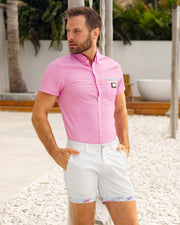 A male model is outdoors wearing the Padam Pink Stretch Shirt, a solid light pink color with blue and pastel yellow stripes on the front pocket. The model completes the look with the White Lotus street chino shorts from DC2 Clothing based in Miami.