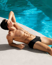 Physique model under the sun in swimming pool wearing the Swim Brief silhouette of the new JET BLACK dark color men's swimsuit by DC2 Miami for BANG menswear Miami.