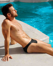 Male model sitting poolside wearing a Swim Sunga  of the new JET BLACK dark color men's swimsuit by DC2 Miami for BANG menswear Miami.