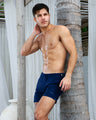 A muscular man with a toned, oiled chest is standing against a palm tree wearing the NAVY BOOMER Tailored Shorts. He is outdoors in a tropical setting, with white curtains and wooden structures in the background. Stylish men's beachwear by BANG! Clothes Miami.