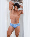 Male model wearing Escapade Swim Brief in blue and white monogram pattern, standing between white curtains, arms raised behind his head.