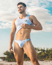 A muscular man models the FORZA WHITE Swim Mini Brief and matching tank top, posing on a sunny beach with palm trees in the background. Designed by BANG! Clothes, a premier men's beachwear brand from Miami, this outfit features a sleek design with Italian-inspired accents, ideal for making a stylish statement at the beach or poolside.